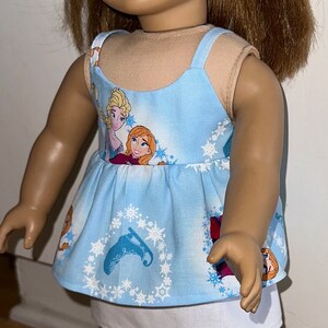 May include: A doll wearing a blue and white dress with a snowflake pattern and a white underskirt. The dress features a print of the characters Elsa and Anna from the movie Frozen. The doll is wearing a blue headband with white snowflakes.