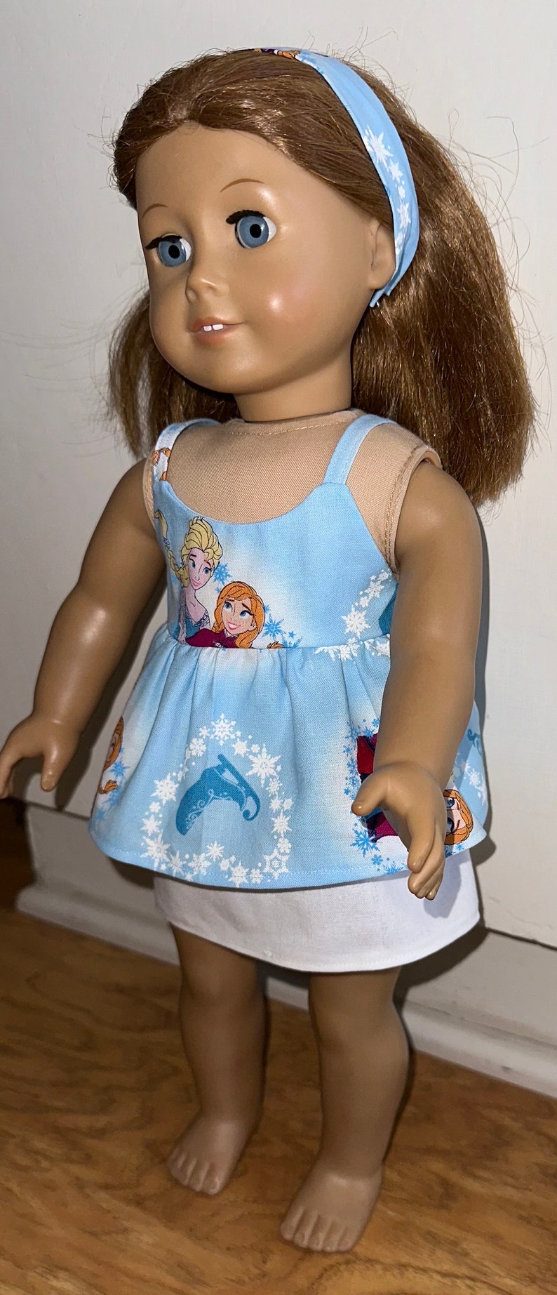 May include: A doll wearing a blue and white dress with a snowflake pattern and a white underskirt. The dress features a print of the characters Elsa and Anna from the movie Frozen. The doll is wearing a blue headband with white snowflakes.