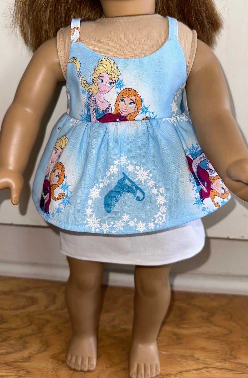 May include: A blue sleeveless dress with a white underskirt. The dress features a pattern of snowflakes and the characters Elsa and Anna from the movie Frozen.