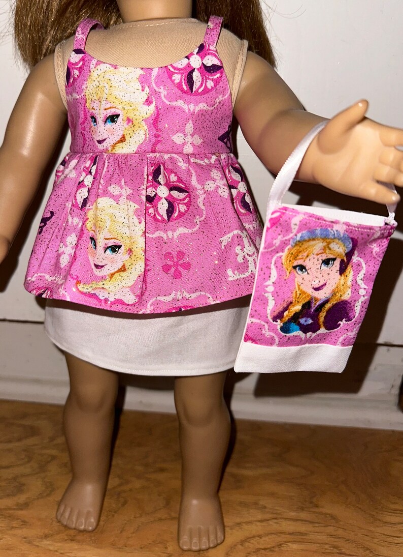 May include: Pink and white doll dress with a glittery pattern featuring a cartoon image of Elsa from Frozen. The dress has a white underskirt and comes with a matching pink and white purse with a cartoon image of Elsa.