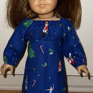 May include: A doll wearing a long-sleeved, blue dress with a festive Christmas print. The dress features illustrations of Christmas trees, nutcrackers, candy canes, and a ballerina. The doll has brown hair and blue eyes.