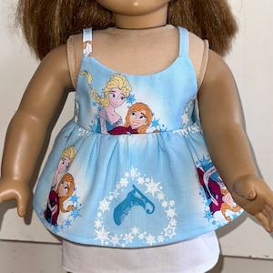 May include: A doll wearing a blue sleeveless dress with a white underskirt. The dress has a pattern of snowflakes and images of the characters from the movie Frozen. The doll has brown hair and blue eyes.
