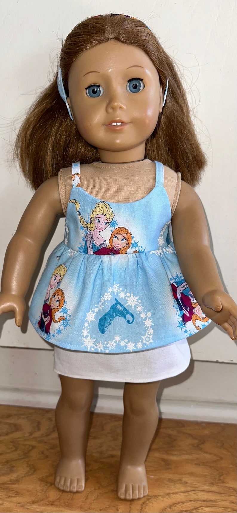 May include: A doll wearing a blue sleeveless dress with a white underskirt. The dress has a pattern of snowflakes and images of the characters from the movie Frozen. The doll has brown hair and blue eyes.