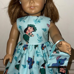 May include: A doll wearing a light blue dress and matching purse, both adorned with cartoon character prints. The dress features a sleeveless design and a flared skirt. The doll has brown hair and blue eyes, with a headband matching the dress.