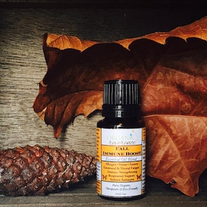 以下が含まれることがあります： 黒いキャップが付いた小さな茶色のガラス瓶で、「ORGANIC reverence FALL IMMUNE BOOST Essential Oil Blend」とラベルが貼られています。瓶は、茶色の葉と松ぼっくりのある木製の表面の上に置かれています。