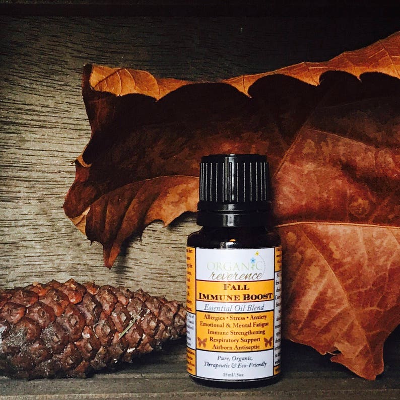 May include: A small brown glass bottle with a black cap, labeled "ORGANIC reverence FALL IMMUNE BOOST Essential Oil Blend". The bottle is sitting on a wooden surface with a brown leaf and a pine cone.
