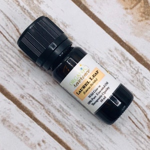 May include: A 10 ml bottle of organic laurel leaf essential oil. The bottle is dark brown glass with a black cap. The label is white with green text and a small yellow flower graphic. The label reads "ORGANIC reverence LAUREL LEAF (Laurus Nobilis) Wild Grown 100% Pure Essential Oil Therapeutic Quality 10 ml"