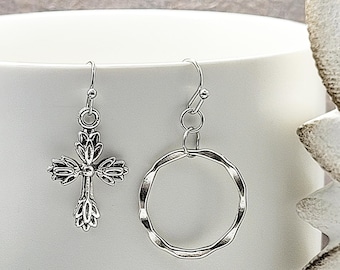 May include: A pair of silver earrings. One earring is a cross with ornate detailing. The other earring is a wavy circle. Both earrings have a hook closure. The earrings are displayed against a white background.