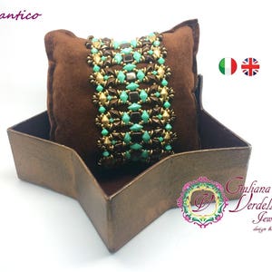Peut inclure: Une boîte en daim marron avec un couvercle en forme d'étoile, contenant un bracelet en perles turquoise et dorées. Le bracelet est composé de petites perles carrées disposées en un motif géométrique. La boîte est étiquetée "Chantico" et "Giuliana Verdelli Jewelry design & creations".