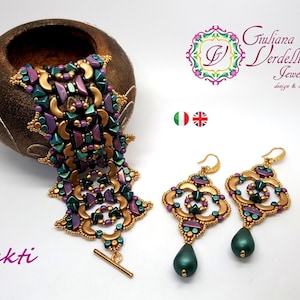 Könnte beinhalten: Ein goldenes, grünes und violettes Perlenarmband und passende Ohrringe mit einem tropfenförmigen Anhänger. Der Schmuck wird auf einer weißen Oberfläche mit einer Holzschale im Hintergrund präsentiert. Der Text "Shakti" ist in Lila unter dem Armband geschrieben.