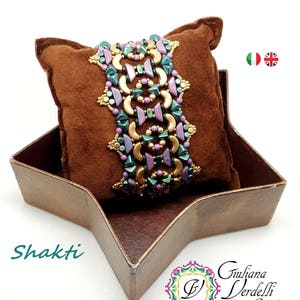 Könnte beinhalten: Ein Armband aus Perlen mit einem geometrischen Muster in Grün-, Lila- und Goldtönen. Das Armband wird auf einem braunen Wildlederkissen in einer sternförmigen Schachtel präsentiert. Der Text "Shakti" ist auf der Schachtel sichtbar. Der Text "Giuliana Verdelli Jewelry design & creations" ist ebenfalls auf der Schachtel sichtbar.