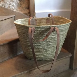 May include: A large, oval-shaped basket made of natural straw with two long, brown leather straps. The basket is sitting on wooden stairs.