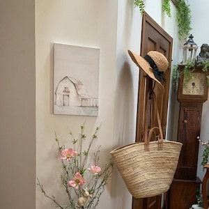 May include: A white farmhouse with a brown roof and a window with a flower box is depicted in a watercolor painting on a canvas. The painting is hanging on a white wall next to a straw hat and a large woven basket.