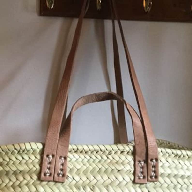 May include: Close-up of the top of a woven straw tote bag with brown leather handles. The handles are stitched with white thread and have metal rivets.