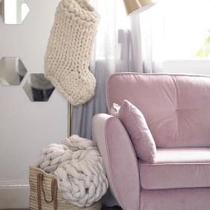 May include: A white knitted stocking hanging on a gold stand, a white knitted throw blanket in a woven basket, and a pink velvet sofa with a white pillow.