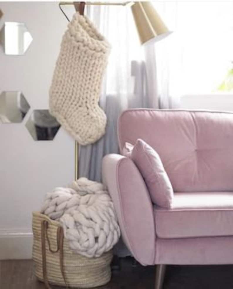 May include: A white knitted stocking hanging on a gold stand, a white knitted throw blanket in a woven basket, and a pink velvet sofa with a white pillow.
