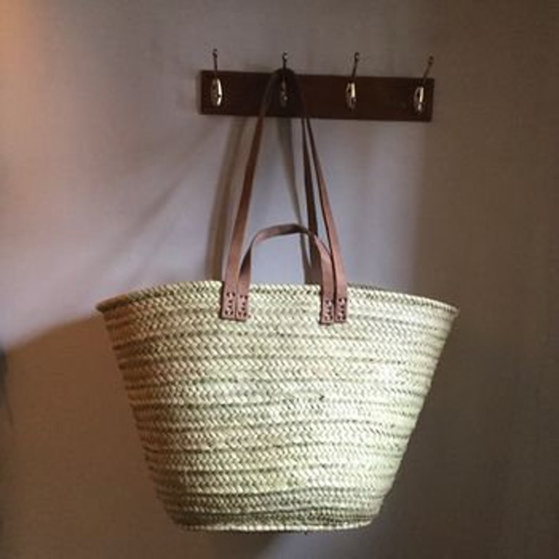 May include: A large, natural woven basket with brown leather handles. The basket is hanging on a hook.