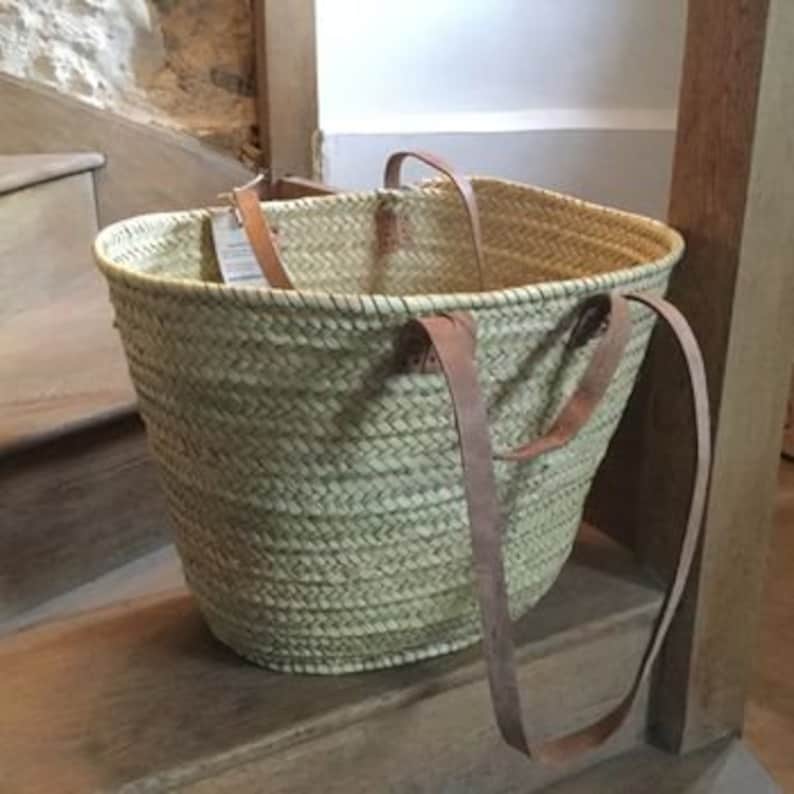 May include: A large, natural woven basket with leather straps. The basket is light brown and has a rounded shape. The straps are long enough to be worn over the shoulder.