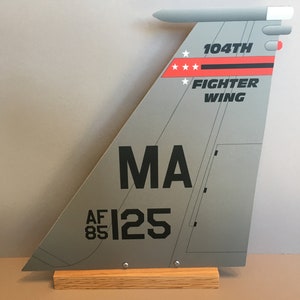 May include: A gray metal model of a fighter jet tail fin with black markings. The tail fin has the text "104th Fighter Wing" and "MA AF85 125" printed on it. The model is mounted on a wooden base.