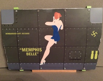 May include: A green, gray, and blue nose art decal featuring a pin-up girl in a blue swimsuit. The decal has the text "Memphis Belle" and "Bombardier Capt. VB Evans".