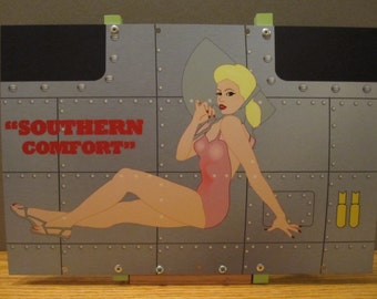Puede incluir: Un letrero de metal con una chica pin-up que lleva un traje de baño rosa y un sombrero blanco. El letrero tiene un fondo gris con remaches y el texto "SOUTHERN COMFORT" en rojo.