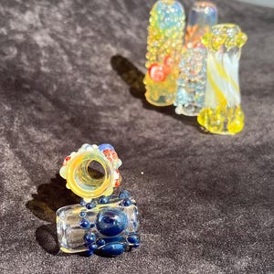 Può includere: Una collezione di opere d'arte in vetro colorato. I pezzi includono un oggetto di vetro trasparente e rettangolare con dettagli di bolle blu e un'apertura circolare, insieme a diverse sculture cilindriche in vetro.