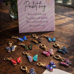 May include: A selection of butterfly charms with gold-coloured clasps, displayed on a wooden surface. A sign reading "Pocket hug" is in the background. The butterflies are in various colours, including blue, red, and purple.