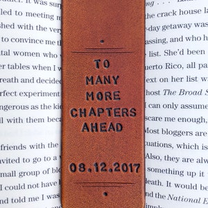 May include: A brown leather bookmark with the embossed text "TO MANY MORE CHAPTERS AHEAD" and the date "08.12.2017". The bookmark is rectangular with a rounded bottom, designed for use in books.