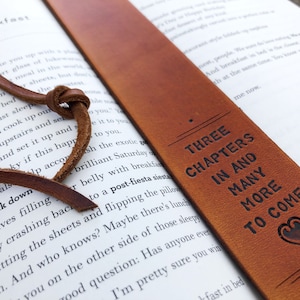 May include: A brown leather bookmark with a knotted tassel and the text "Three Chapters In And Many More To Come" with a heart symbol.
