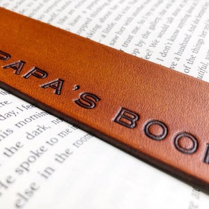 May include: A brown leather bookmark with the embossed text "PAPA'S BOOK". The bookmark is rectangular and is placed on top of a book with printed text. The leather has a textured surface.