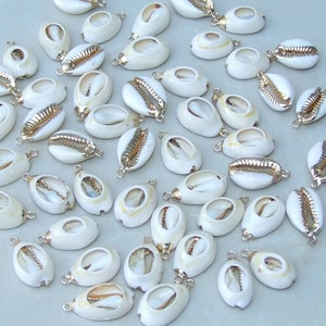 May include: A collection of white cowrie shells with gold-colored accents and small silver-colored loops. These shells are a natural off-white color and are arranged on a light gray surface. The shells are a popular choice for jewelry making and crafts.