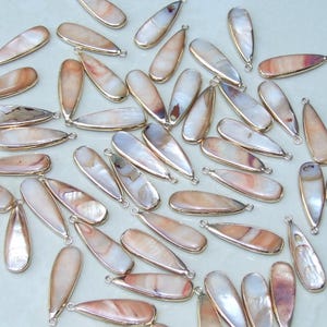 Op de afbeelding: Een verzameling van 50 parelmoer witte en bruine schelp teardrop hangers met goudkleurige springringen. De hangers zijn ongeveer 3,8 cm lang.