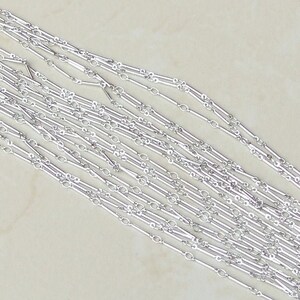 May include: A close-up of a silver chain necklace with a unique design featuring alternating oval and rectangular links. The chain is laid out in a fan shape, showcasing the intricate details of the design.