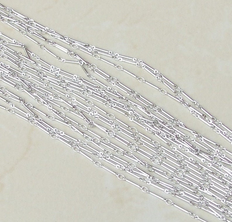 May include: A close-up of a silver chain necklace with a unique design featuring alternating oval and rectangular links. The chain is laid out in a fan shape, showcasing the intricate details of the design.