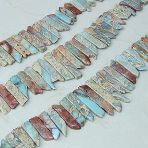 May include: A strand of turquoise, brown, and beige gemstone beads. The beads are long and narrow, with a jagged edge. The beads are arranged in a staggered pattern.