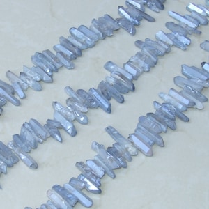 May include: A strand of light blue crystal points, also known as quartz points, arranged in rows on a white surface. The points are various lengths and have a shimmery, iridescent finish.