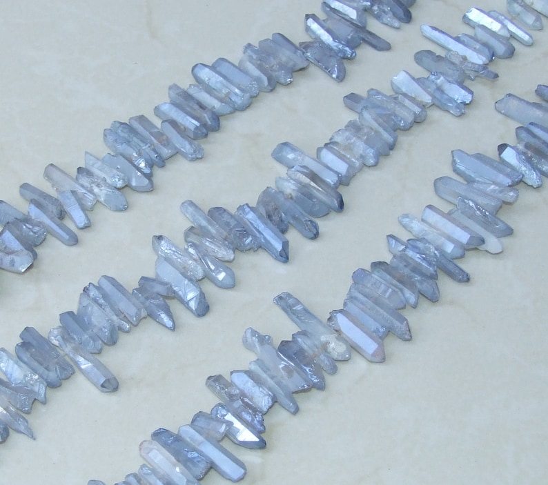 May include: A strand of light blue crystal points, also known as quartz points, arranged in rows on a white surface. The points are various lengths and have a shimmery, iridescent finish.