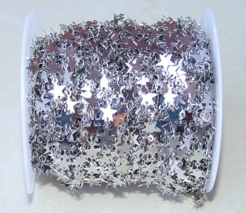 May include: A spool of silver star chain. The chain is made of small, flat, silver stars that are linked together. The chain is wound around a white spool.