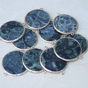 May include: A collection of round, dark green and blue gemstone pendants with gold-colored rims. Each pendant has a unique marbled pattern and small loops for attaching to jewelry. The pendants are arranged on a light-colored surface.
