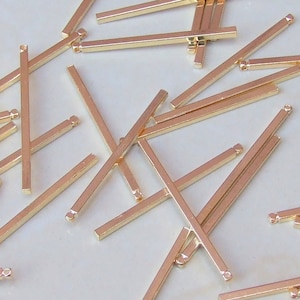 Puede incluir: Una colección de pequeñas barras metálicas rectangulares de color oro rosa dispersas sobre una superficie. Cada barra tiene un pequeño bucle en un extremo, lo que sugiere que son componentes para la fabricación de joyas o manualidades similares.