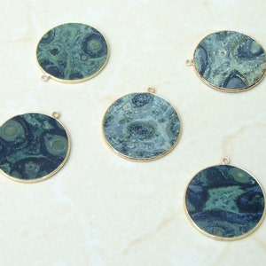 May include: Five round, flat pendants with a dark green and blue marbled pattern. Each pendant has a gold-colored metal rim and a small loop at the top for attaching to a chain or other jewelry component. The pendants are displayed on a light-colored surface.