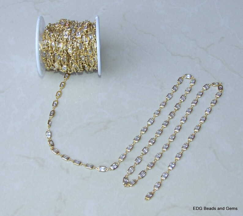 Op de afbeelding: Een spoel met een gouden ketting met heldere rechthoekige stenen. De ketting ligt in een lange lijn op een wit oppervlak.