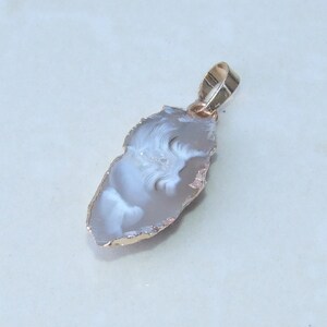 May include: A pendant featuring a polished, irregularly shaped agate stone with a gold-colored metal bail. The agate has a light gray and white swirling pattern with a gold-colored edge.