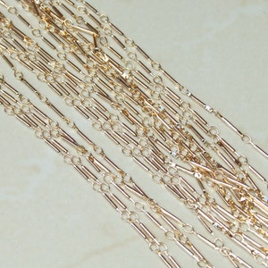 May include: A close-up of multiple strands of gold chain with a bar link design. The chain is made of small, thin, gold-colored metal bars connected by small, round, gold-colored links.