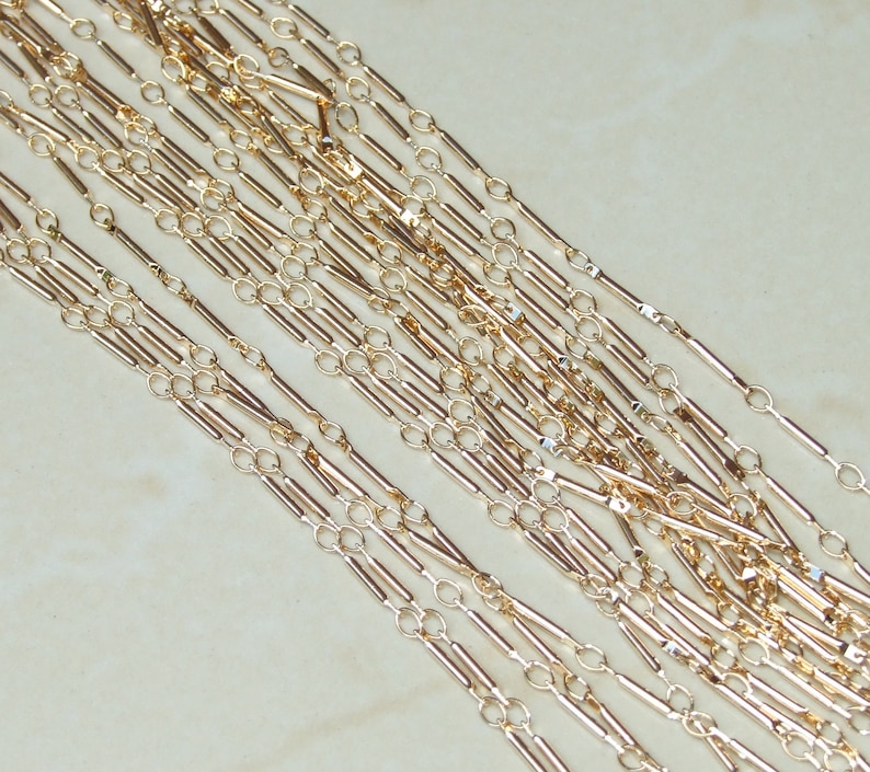 May include: A close-up of multiple strands of gold chain with a bar link design. The chain is made of small, thin, gold-colored metal bars connected by small, round, gold-colored links.