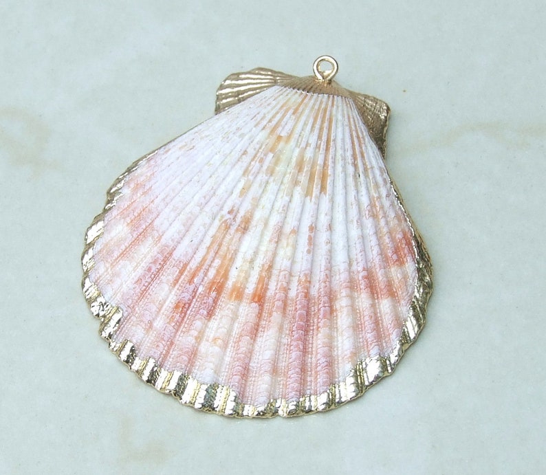Op de afbeelding: Een grote, sint-jakobsschelpvormige hanger met een goudkleurige rand. De schelp heeft een natuurlijk patroon van witte, roze en oranje tinten. Een kleine gouden lus is aan de bovenkant bevestigd om op te hangen. Ideaal voor het maken van sieraden of kustdecoratie.