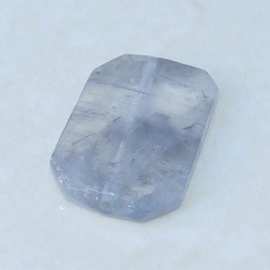 May include: A translucent, light blue gemstone bead with a rectangular shape and beveled edges. The bead has a hole drilled through it, suggesting it is for jewelry making or crafting. The stone has a subtle, cloudy appearance.