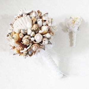 May include: A white seashell bridal bouquet with a smaller matching boutonniere. The bouquet is made of various sizes and shapes of seashells, tied together with white ribbon.