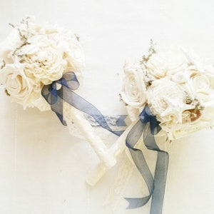 May include: Two ivory-colored bridal bouquets with a variety of flower shapes and textures. Each bouquet is accented with a navy blue ribbon and lace. The stems are wrapped in ivory lace. The bouquets are arranged on a white surface.