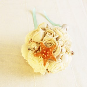 May include: A bouquet of white silk roses and seashells with a starfish in the center. The bouquet is tied with a light blue ribbon.